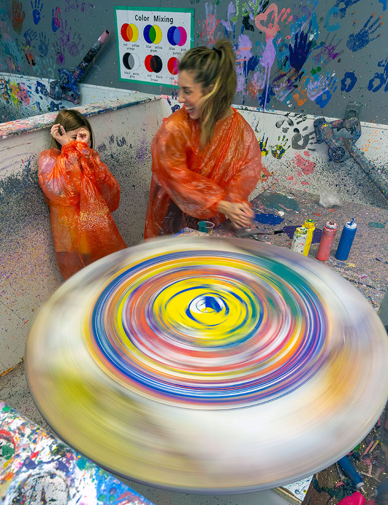 Raleigh 8-year-old Coletter Luwisch covers her face from splatter as mom Lauren spins the wheel that holds their artwork.